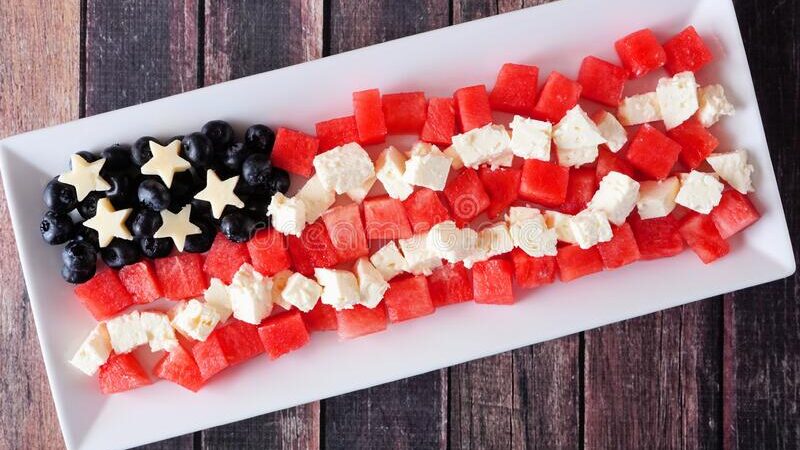 A rectangular platter with blueberries, star-shaped cheese slices, watermelon cubes, and white cheese cubes arranged to resemble the American flag on a wooden background.