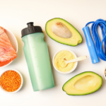 A flat lay of healthy foods and fitness items: a water bottle, salmon fillet, avocado halves, lentils, chickpeas, protein powder with a scoop, and a blue jump rope on a white background.