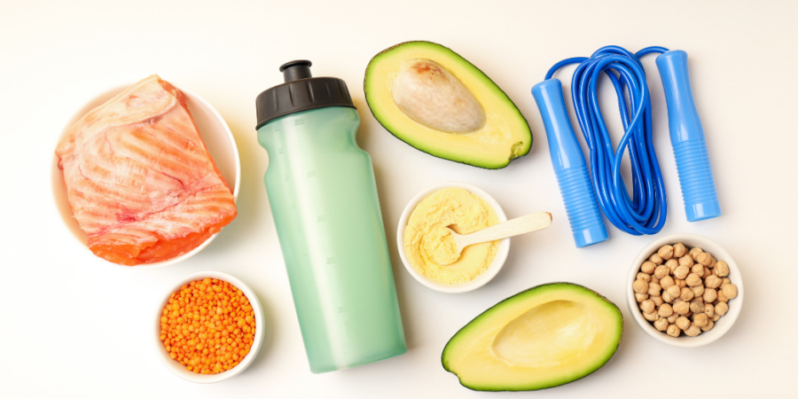 A flat lay of healthy foods and fitness items: a water bottle, salmon fillet, avocado halves, lentils, chickpeas, protein powder with a scoop, and a blue jump rope on a white background.