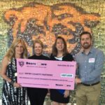 Four people stand smiling and holding a large pink ceremonial check for $157,500 made out to the BBFWF Charity Partners from Bears Care, with a photo collage wall in the background.