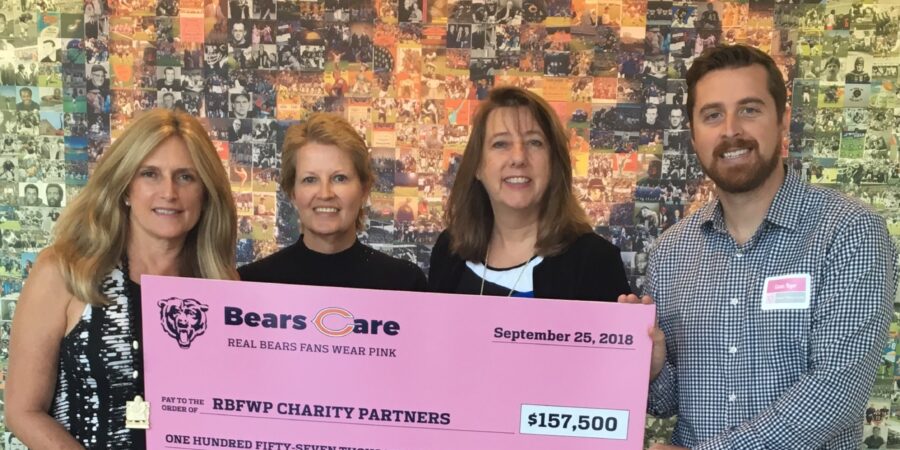 Four people stand smiling and holding a large pink ceremonial check for $157,500 made out to the BBFWF Charity Partners from Bears Care, with a photo collage wall in the background.