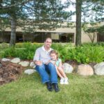 A man and a young girl sit on grass under trees, smiling, with a brick building and landscaped bushes and rocks in the background.