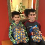 Two boys sit on a bench in front of a red wall and mirror, smiling and holding blue drawstring bags with yellow emoji faces printed on them.