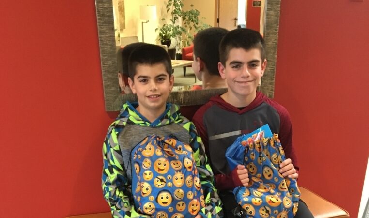 Two boys sit on a bench in front of a red wall and mirror, smiling and holding blue drawstring bags with yellow emoji faces printed on them.