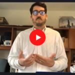 A man wearing glasses and a white shirt speaks during a video call, gesturing with his hands. He stands in an office with wooden shelves, pictures, and various items in the background. The Zoom logo appears in the corner.