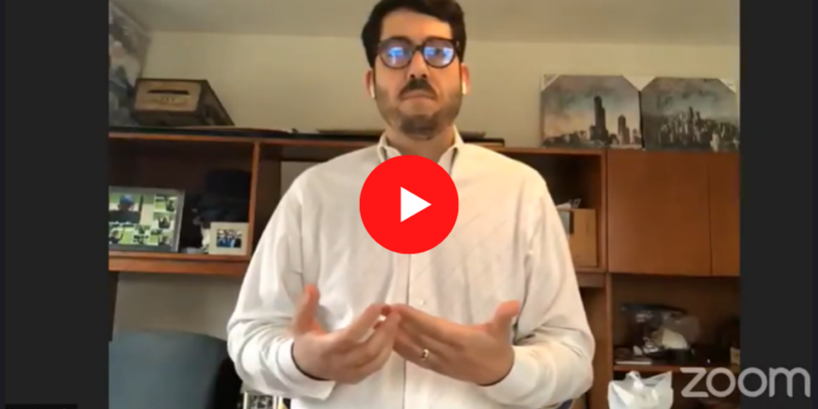 A man wearing glasses and a white shirt speaks during a video call, gesturing with his hands. He stands in an office with wooden shelves, pictures, and various items in the background. The Zoom logo appears in the corner.