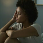 A woman sits on a bed with her knees pulled to her chest, resting her head on her hand and looking thoughtfully into the distance. A lit lamp and striped pillow are visible in the background.