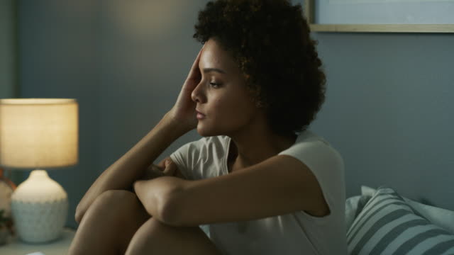 A woman sits on a bed with her knees pulled to her chest, resting her head on her hand and looking thoughtfully into the distance. A lit lamp and striped pillow are visible in the background.