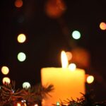 A lit candle surrounded by pine branches with colorful, blurred holiday lights glowing softly in the dark background.