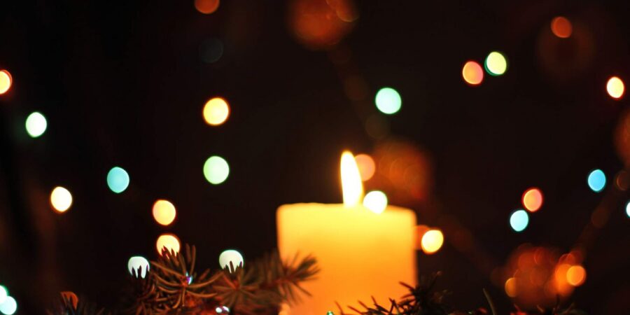 A lit candle surrounded by pine branches with colorful, blurred holiday lights glowing softly in the dark background.