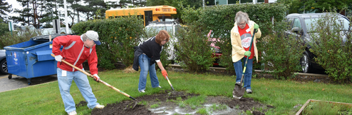 Three people use shovels to work on a garden bed near parked cars, a school bus, and a blue dumpster, with bushes in the background.