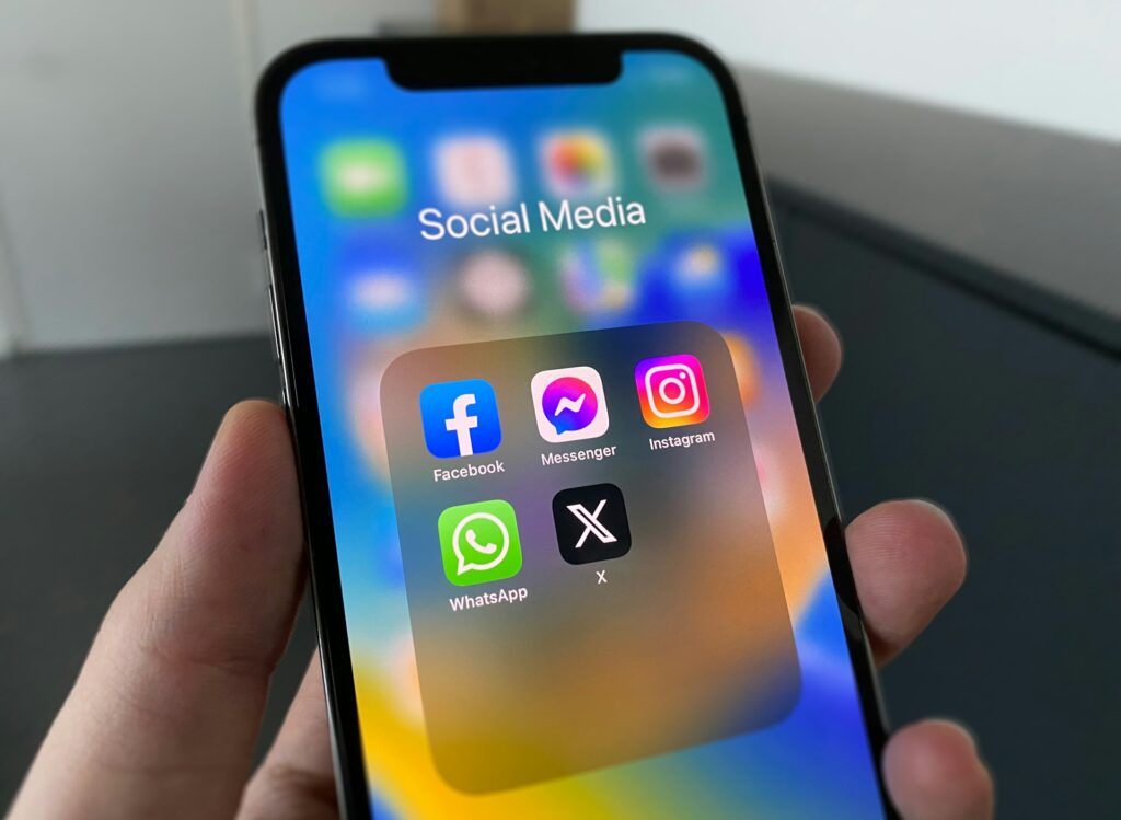 A hand holds a smartphone displaying a folder labeled "Social Media" with icons for Facebook, Messenger, Instagram, WhatsApp, and X on the screen. The background is blurred.