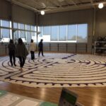 Four people walk barefoot on a large, circular labyrinth mat laid out on a gymnasium floor, with exercise balls, windows, and gym equipment visible in the background.
