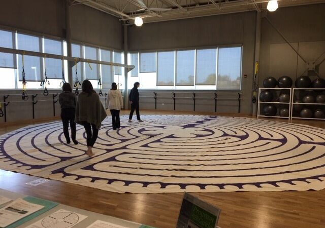 Four people walk barefoot on a large, circular labyrinth mat laid out on a gymnasium floor, with exercise balls, windows, and gym equipment visible in the background.