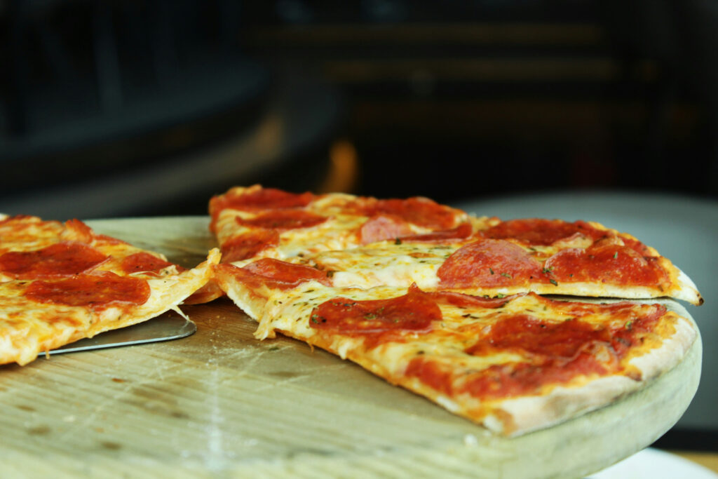 A thin-crust pepperoni pizza with several slices cut sits on a wooden pizza board; one slice is being lifted slightly with a metal spatula.