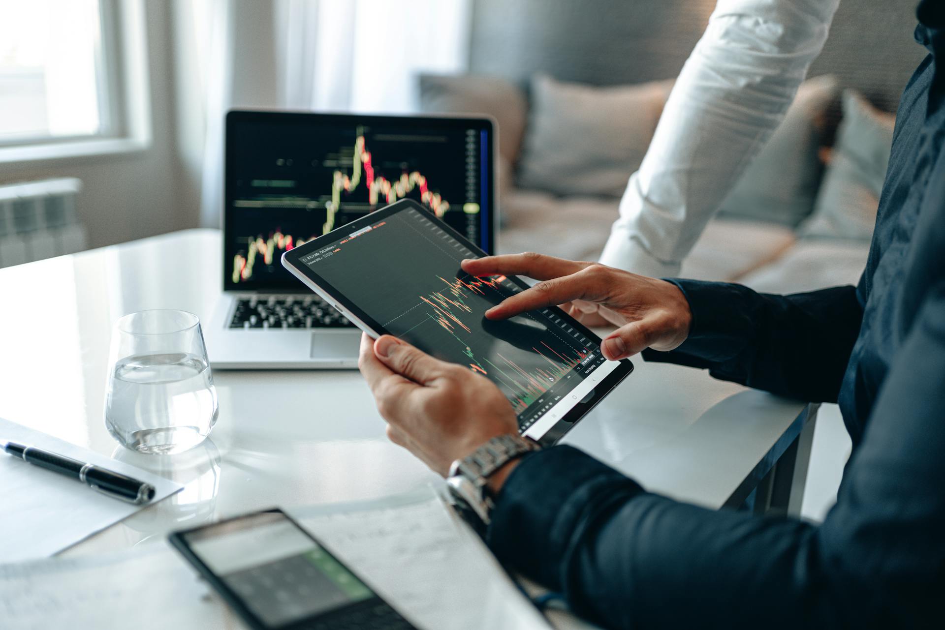 A person holds a tablet displaying stock market charts, with a laptop showing similar graphs in the background. A glass of water, a smartphone, and papers are on the table. Another person stands nearby.