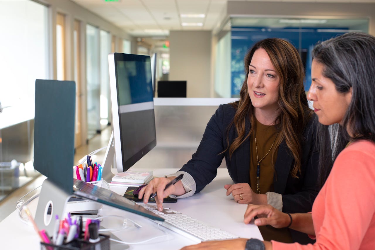 Two women sit at a desk in an office, looking at a computer monitor. One woman points at the screen with a pen while the other uses the keyboard. Office supplies and computer accessories are on the desk.