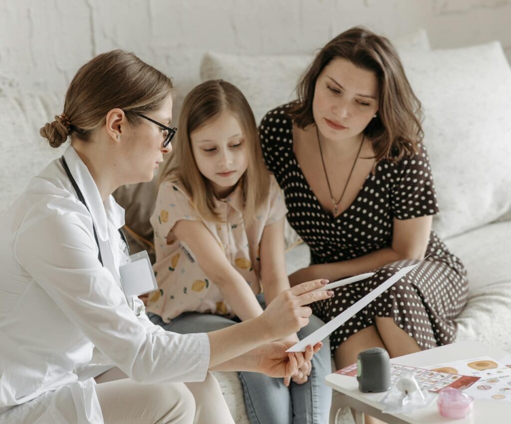 A doctor shows medical documents to a young girl and her mother, who are sitting together on a couch, attentively listening and looking at the papers. Medical equipment is visible on the table in front of them.