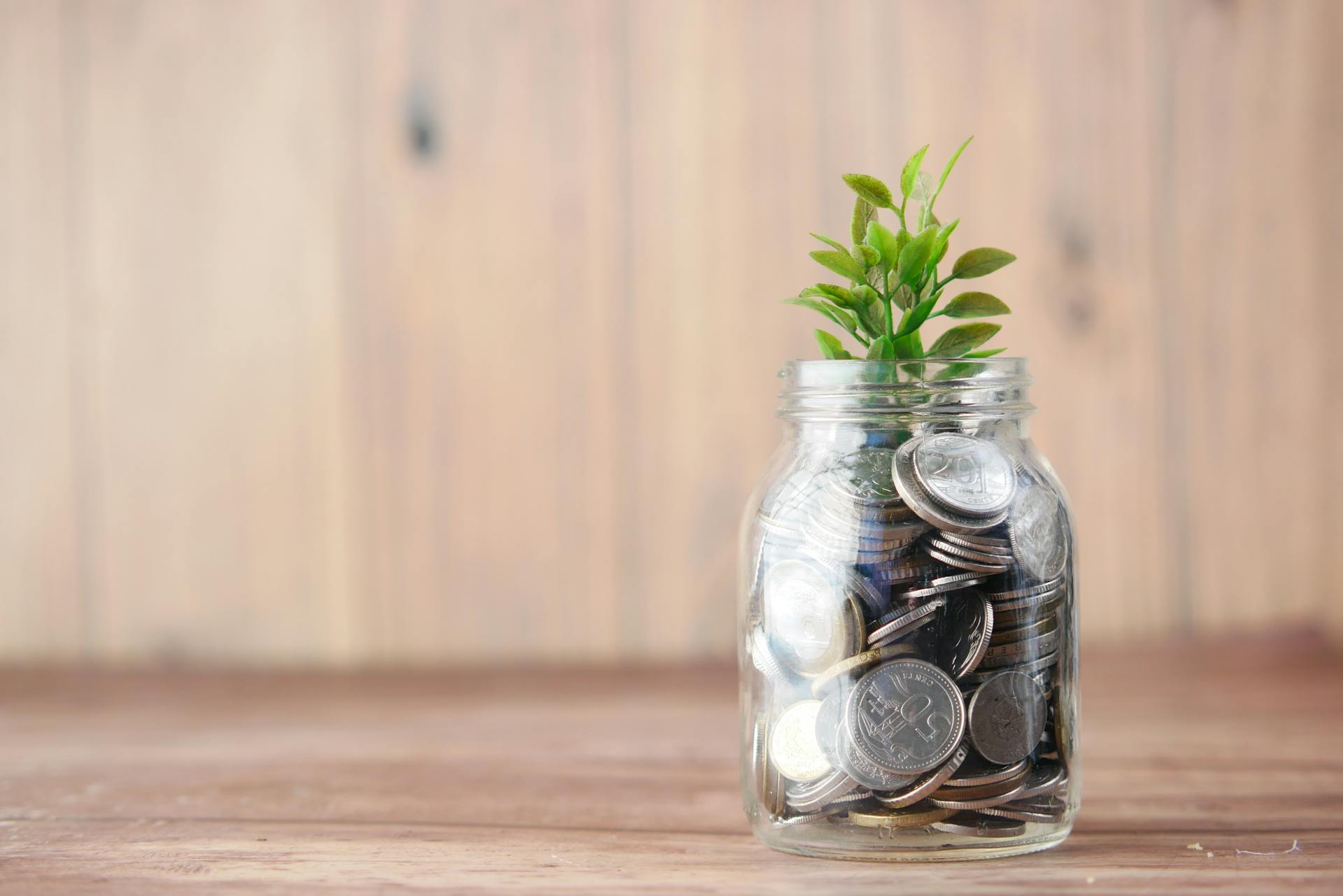 A glass jar filled with coins sits on a wooden surface, with a small green plant growing from the top, symbolizing financial growth and saving. The background is light wood paneling.