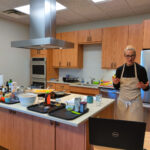 A person in an apron stands in a modern kitchen, speaking and gesturing toward a laptop on the counter, surrounded by various cooking ingredients and utensils.