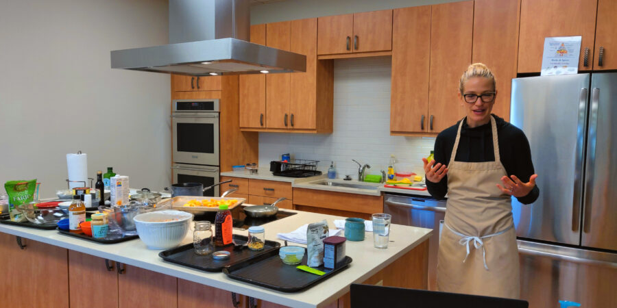 A person in an apron stands in a modern kitchen, speaking and gesturing toward a laptop on the counter, surrounded by various cooking ingredients and utensils.