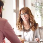 A woman with glasses and wavy hair smiles while talking to a man across a table in a bright café. She wears a statement necklace and light-colored clothing, holding a tablet with a coffee and pastries on the table.