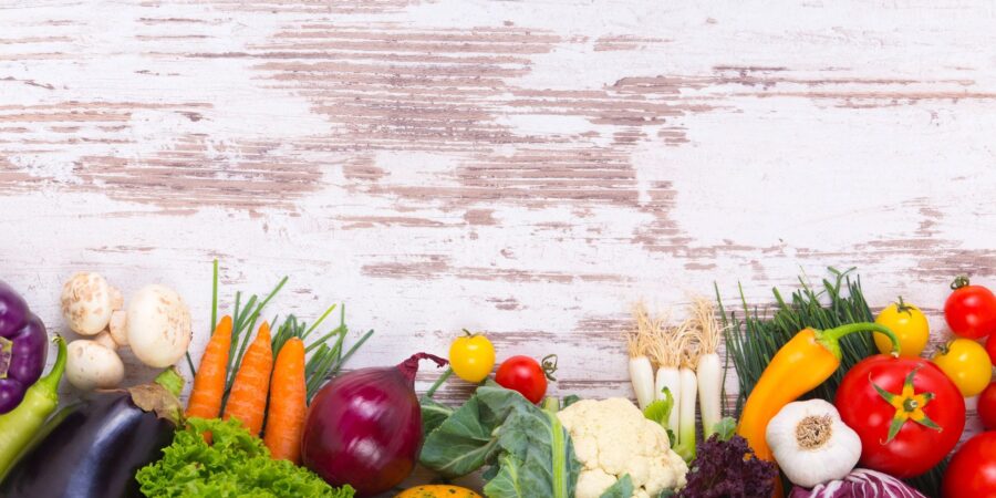 A colorful variety of fresh vegetables, including eggplant, carrots, lettuce, onion, tomatoes, garlic, cauliflower, and peppers, arranged in a row at the bottom against a rustic white wooden background.