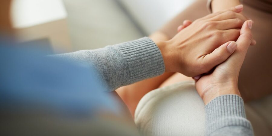Close-up of one person gently holding another person's hands, offering comfort and support. Both are wearing sweaters, and the scene conveys empathy and compassion.