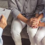 A couple sits close together on a couch, holding hands for support, while a person across from them takes notes on a clipboard, suggesting a counseling or therapy session.