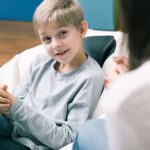 A young boy with light brown hair sits on a beanbag chair, smiling and talking with an adult whose face is out of focus in the foreground. The setting appears to be casual and comfortable indoors.