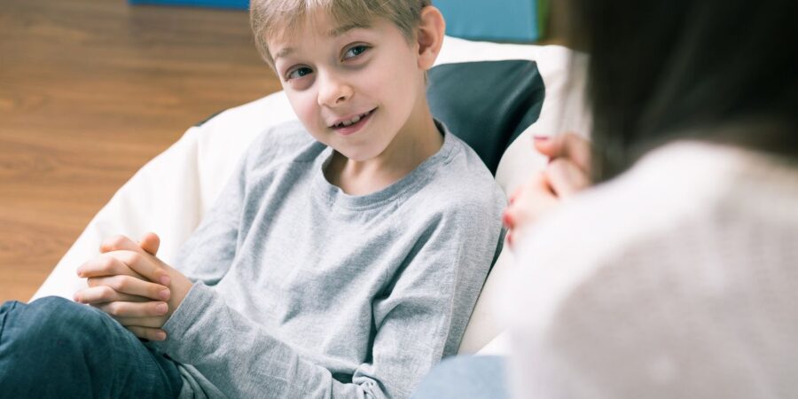 A young boy with light brown hair sits on a beanbag chair, smiling and talking with an adult whose face is out of focus in the foreground. The setting appears to be casual and comfortable indoors.
