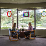 Two women sit facing each other in chairs by a large window with colorful stained glass panes, talking in a bright room with trees and greenery visible outside.