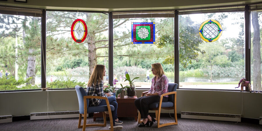 Two women sit facing each other in chairs by a large window with colorful stained glass panes, talking in a bright room with trees and greenery visible outside.