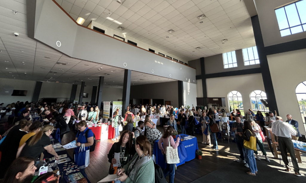 A large group of people attend an indoor event with booths and tables set up, where attendees are interacting, browsing materials, and engaging with exhibitors in a spacious room with tall windows and high ceilings.