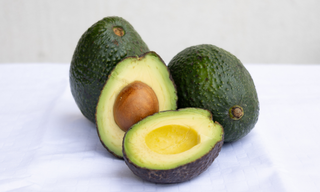 Three avocados on a white surface: one whole, one halved with the pit, and one halved without the pit, showing the green flesh and bumpy dark green skin.