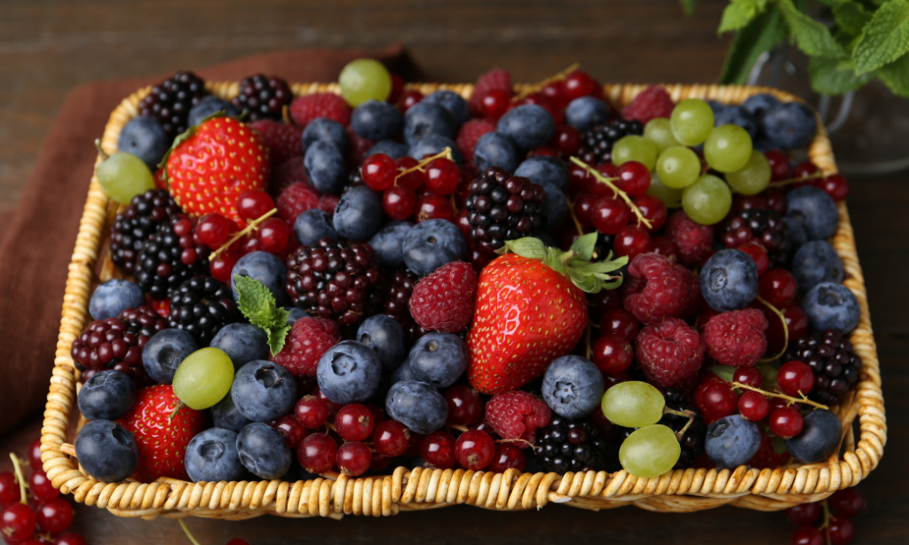 A wicker basket filled with fresh mixed berries and grapes, including strawberries, blueberries, raspberries, blackberries, red currants, and green grapes, set on a dark wooden surface.