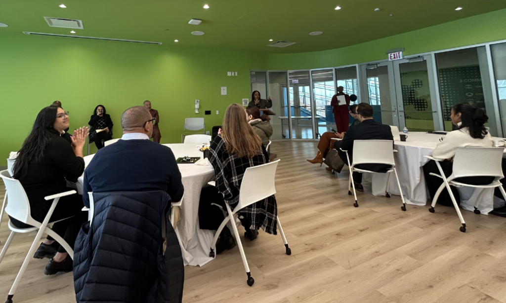 People sit around round tables in a bright room with green walls, listening to two speakers standing at the front. The atmosphere is casual and collaborative, with natural light from large windows.