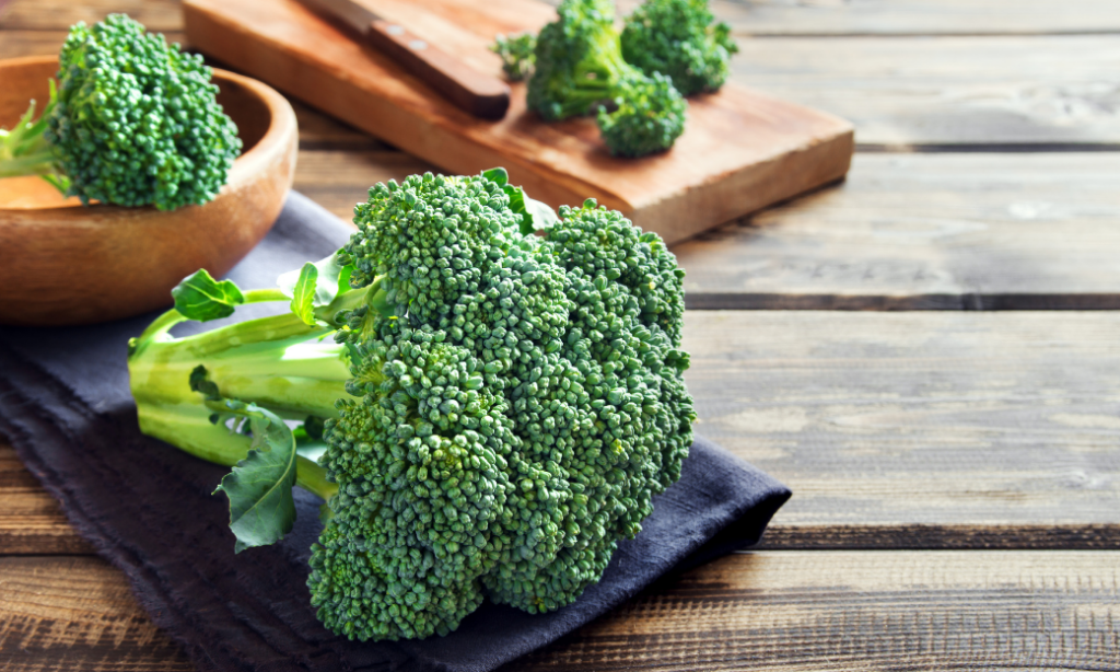 A fresh head of broccoli rests on a dark cloth on a wooden table, with additional broccoli pieces in a wooden bowl and on a cutting board in the background.
