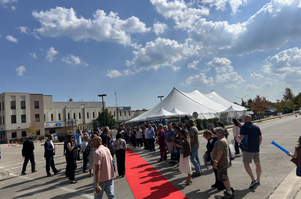 A crowd of people stands in line along a red carpet outside a large white building and tent on a sunny day with blue skies and scattered clouds.
