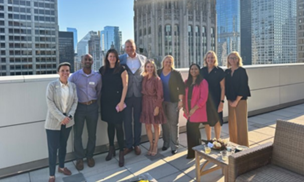 A group of nine people stand smiling on a rooftop terrace in a city, with tall buildings in the background on a sunny day. There is patio furniture and a small table with food in the foreground.