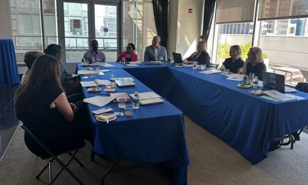 Eight people sit around a U-shaped table covered with blue tablecloths, engaged in a meeting with papers, laptops, and drinks on the tables. Large windows in the background show a cityscape.