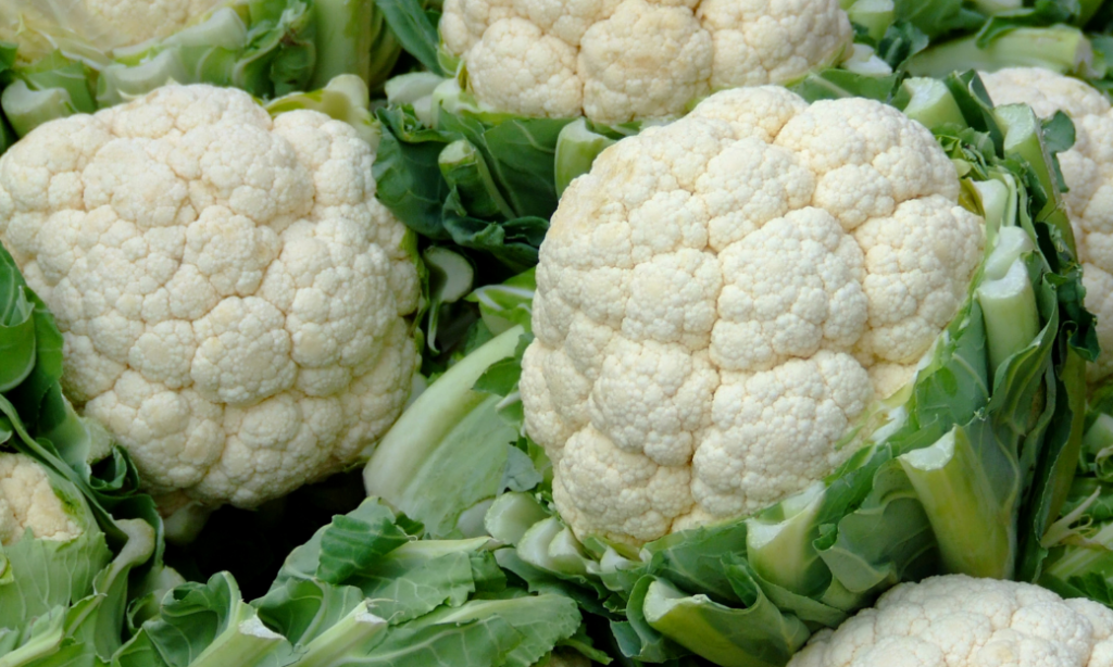 Close-up of several fresh cauliflower heads with white florets and green leaves, piled together.