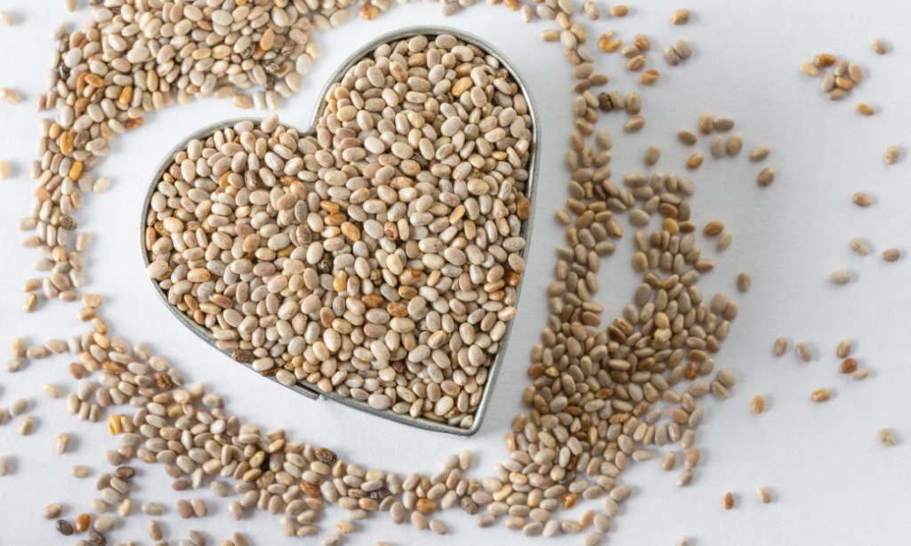 A heart-shaped metal container filled with chia seeds sits on a white surface, with chia seeds scattered around it.