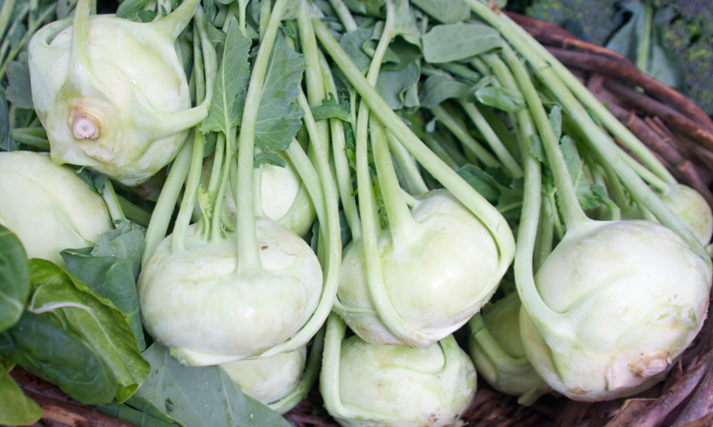 A close-up of several fresh, light green kohlrabi bulbs with stems and leaves, displayed in a woven basket.
