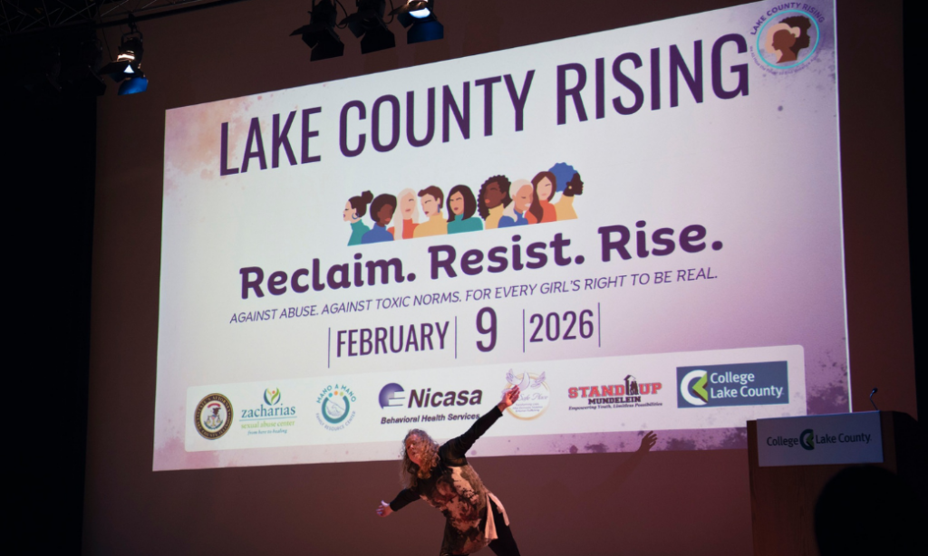 A person gestures on stage in front of a large screen displaying “Lake County Rising: Reclaim. Resist. Rise. Against abuse, against toxic norms. For every girl’s right to be real. February 9, 2026.” Logos of sponsors are below.