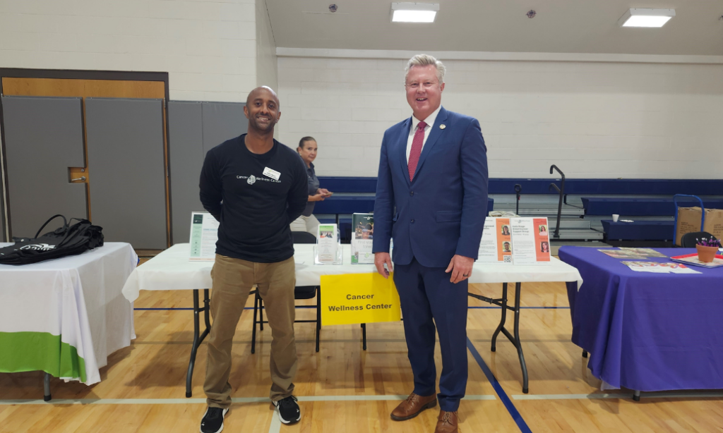 Two men stand and smile in front of a Cancer Wellness Center table at an indoor event. One man wears a black shirt and khaki pants; the other wears a blue suit and tie. Display tables and brochures are visible behind them.