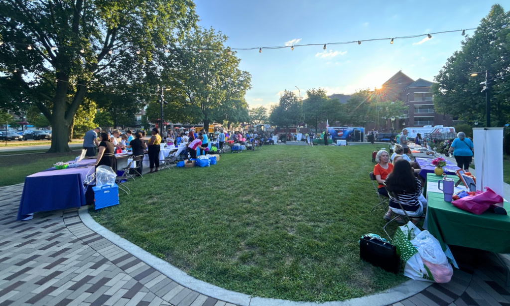A community fair in a park with booths, tables, and people gathered on the grass under string lights, surrounded by trees and buildings in the background during sunset.