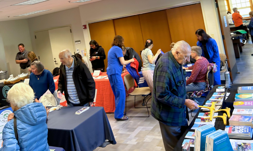 People browse tables with books, pamphlets, and informational materials at a busy indoor event. Some are seated behind tables, others look at displays. The room has bright lighting and several posters on the walls.