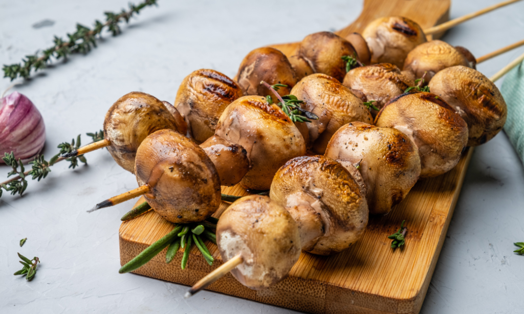 Grilled mushroom skewers arranged on a wooden cutting board, garnished with fresh herbs, with a garlic bulb and sprigs of thyme nearby on a light surface.