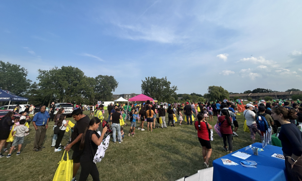 A large crowd gathers outdoors at a community event, with people walking, talking, and holding bags. Several tents and tables are set up on the grass under a clear blue sky with trees in the background.